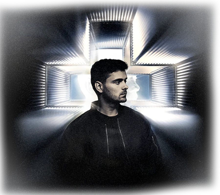Martin Garrix con chaqueta oscura frente a una estructura de luz en forma de cruz.
