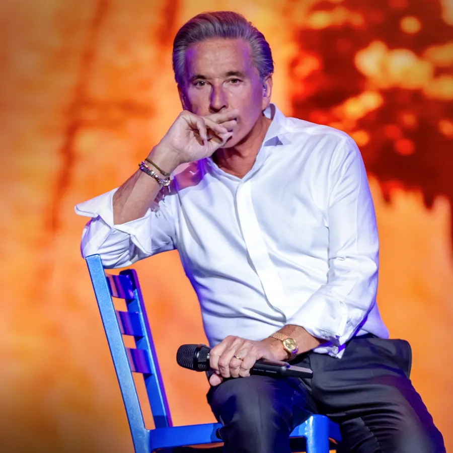 Ricardo Montaner en concierto, sentado en una silla azul con el micrófono en la mano.