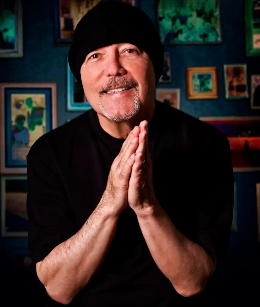 Rubén Blades sonríe con las manos juntas, con gorro negro, frente a una pared llena de cuadros.
