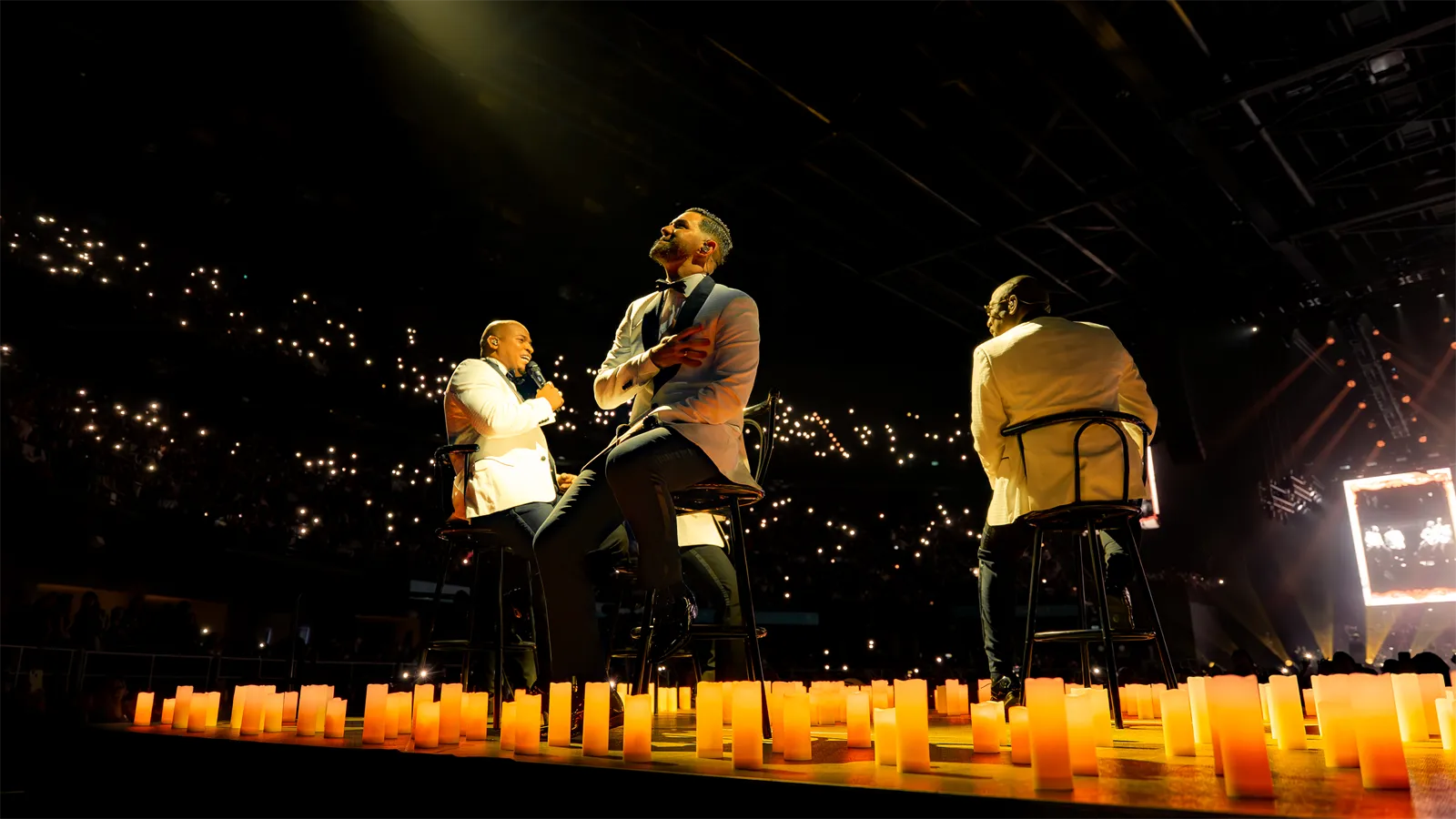 Tres cantantes en el escenario frente a las velas y las luces del público.
