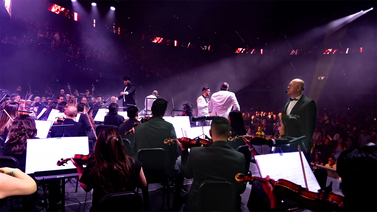 Músicos de la orquesta y cantantes en el escenario con luces moradas.